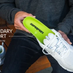 Person holds a lime-green Superfeet insole beside a white athletic shoe, highlighting supportive footwear.”