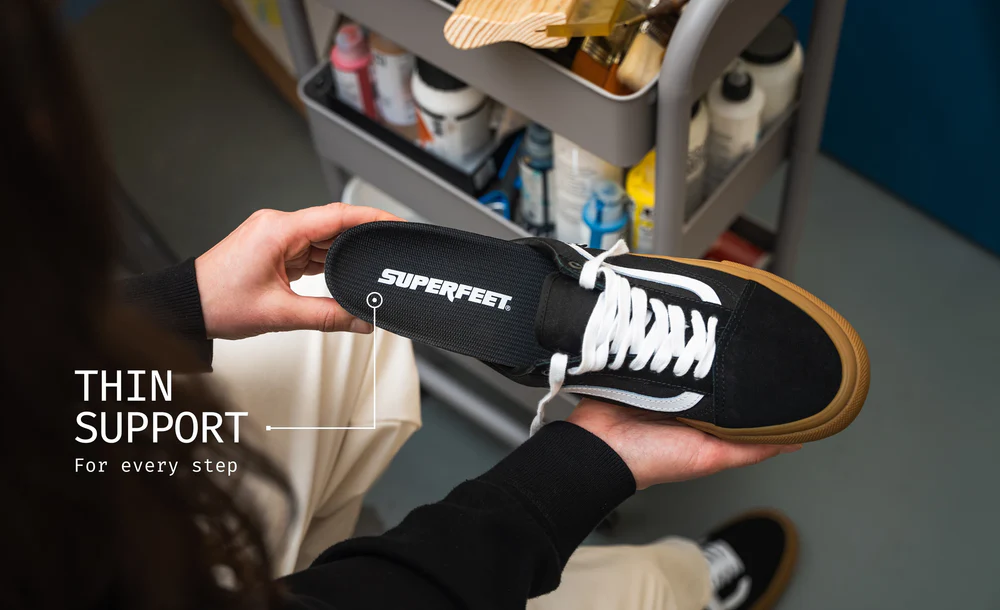 View2_All-Purpose_Support_Low-Arch_2731x1667_f0aaf816-4493-4934-992a-dccaf5797837 - Horizon Industries Close-up of a black Superfeet sneaker being held, with a 'Thin Support' annotation and 'For every step' text in a workshop setting