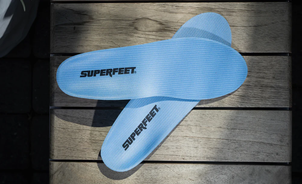View8_All-Purpose_Support_Medium-Arch_2731x1667_420e7a4e-0ad6-48b2-a51d-6fa8b13fa45b - Horizon Industries Light blue Superfeet shoe insoles laid crossing on a wooden bench surface.